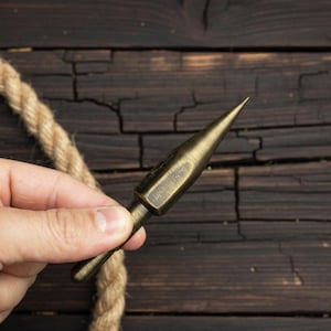 Op de afbeelding: Een bronzen metalen priem met een scherpe, conische punt. De priem wordt in een hand gehouden, met een touw en een donkere houten achtergrond. Het ontwerp van de priem heeft een cilindrische handgreep en een puntige punt, geschikt voor het doorboren of markeren van materialen.