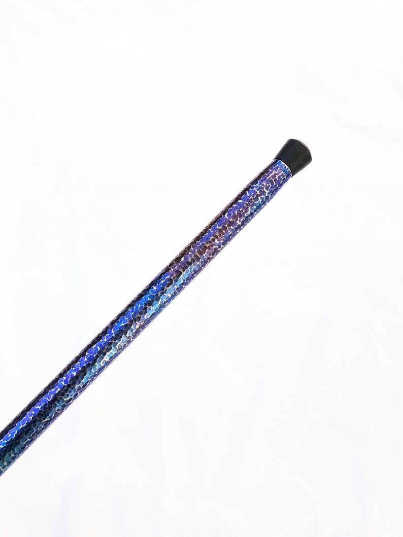 Op de afbeelding: Een lange, cilindrische staaf met een levendige, iriserende afwerking met een herhalend patroon van kleine, glinsterende blauwe, paarse en donker metallic schubben. Het uiteinde van de staaf is afgewerkt met een gladde, zwarte dop.