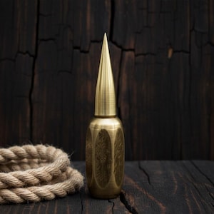 Op de afbeelding: Een messingkleurige, decoratieve fles met een puntige top en gegraveerde bloempatronen. De fles staat tegen een donkere houten achtergrond met een opgerold touw op de voorgrond. Het ontwerp suggereert een vintage esthetiek.