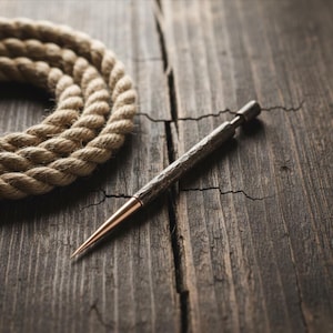 May include: A close-up shot of a textured metal pen with a copper-colored tip, lying on a weathered wooden surface. A coiled rope is in the background. The pen has a hammered finish, suggesting a handcrafted design.