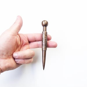 Op de afbeelding: Een bronskleurig metalen gereedschap met een bolvormige bovenkant, een centraal cilindrisch gedeelte met een getextureerde, gehamerde afwerking en een gladde, taps toelopende punt. Het object is gemaakt van een metaalachtig materiaal.