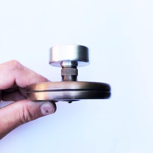 May include: A metal bottle stopper with a silver top and a bronze-coloured base. The stopper has a textured, knurled section and a flat, circular base. The design is simple and elegant.