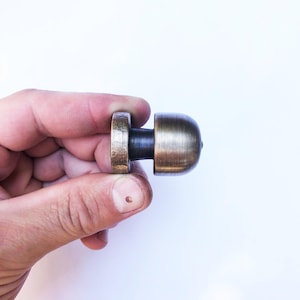 May include: A bronze-colored cabinet knob with a rounded top and a cylindrical base. The knob is held in a hand against a white background. The metal has a slightly aged appearance.