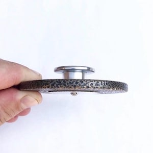 May include: A round, black and silver, metal bottle stopper with a textured surface.