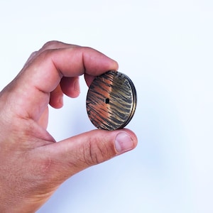 Può includere: Una manopola circolare in metallo caratterizzata da una finitura testurizzata, scura e color rame, con una serie di linee verticali incise sulla superficie. La manopola presenta un piccolo foro esagonale centrale e un bordo liscio e scuro.