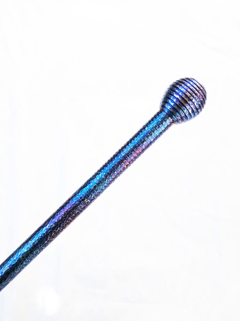 Op de afbeelding: Een lange, slanke metalen staaf met een getextureerd, gehamerd oppervlak met een iriserende blauwe en paarse afwerking. De staaf eindigt in een bolvormige, geribbelde knop aan de bovenkant, met dezelfde metallic uitstraling en kleurverloop.