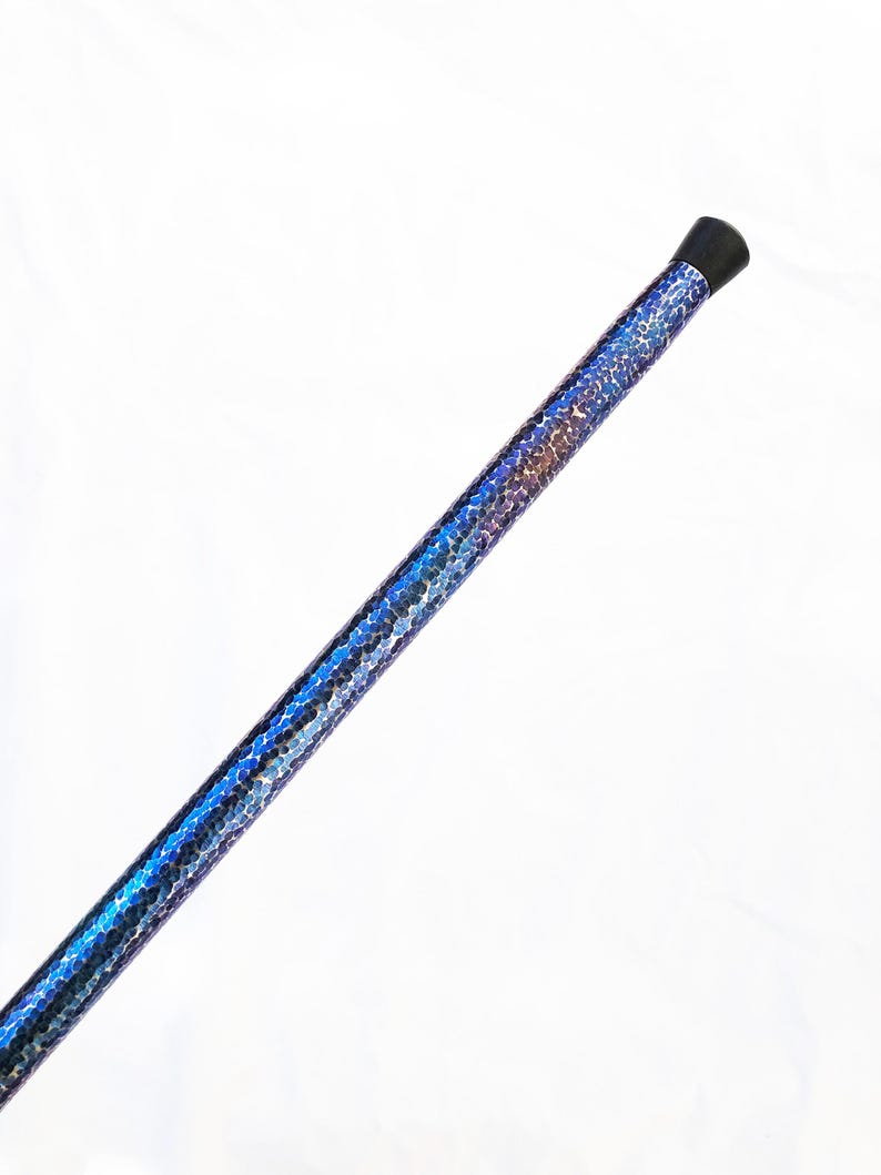 Op de afbeelding: Een lange, cilindrische wandelstok met een levendig, iriserend blauw en paars schubachtig patroon dat de gehele schacht bedekt. De bovenkant van de stok is afgewerkt met een gladde, effen zwarte dop, wat zorgt voor een strak contrast.