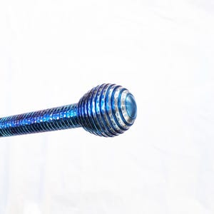 Op de afbeelding: Een metalen gereedschap met een levendige blauwe iriserende afwerking, voorzien van een lange, slanke steel met een spiraalvormige groeftextuur die leidt naar een rond, bolvormig uiteinde met concentrische ribbels en een glad middelpunt.
