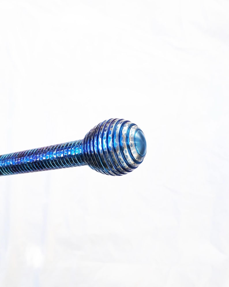 Op de afbeelding: Een metalen gereedschap met een levendige blauwe iriserende afwerking, voorzien van een lange, slanke steel met een spiraalvormige groeftextuur die leidt naar een rond, bolvormig uiteinde met concentrische ribbels en een glad middelpunt.