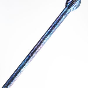 Op de afbeelding: Een lange, slanke metalen staaf met een glinsterende, iriserende blauwe en paarse afwerking. Het oppervlak heeft een fijne, gehamerde textuur over de gehele lengte van de schacht. Het is bekroond met een bolvormige, geribbelde knop.