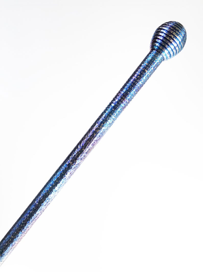Op de afbeelding: Een lange, slanke metalen staaf met een glinsterende, iriserende blauwe en paarse afwerking. Het oppervlak heeft een fijne, gehamerde textuur over de gehele lengte van de schacht. Het is bekroond met een bolvormige, geribbelde knop.
