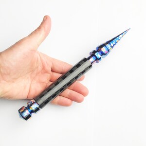 May include: A multi-coloured, pointed tool with a black chequered handle. The tip is a vibrant mix of blues and purples, with a metallic sheen. The handle has a dark, textured pattern. The tool is held against a white background.