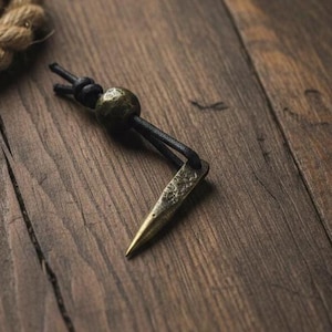 Op de afbeelding: Een handgemaakte messing hanger met een puntig uiteinde, bevestigd aan een zwart koord en een decoratieve kraal. De hanger wordt gepresenteerd op een houten oppervlak, met een touw op de achtergrond. De hanger is ongeveer 5 cm lang.
