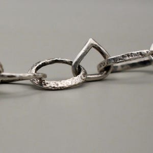 Pode incluir: Close-up de uma corrente de prata com elos texturizados. A corrente apresenta uma variedade de formas de elos, incluindo designs ovais e em forma de gota. O metal tem uma aparência ligeiramente martelada, dando-lhe um toque rústico.