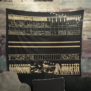 May include: Black tapestry with gold hieroglyphics and figures depicting a procession of people and animals. The tapestry is hanging on a wall with string lights.