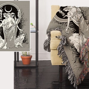 May include: A woven tapestry throw blanket features a black and white illustration of three women standing in a forest with wolves. The women are wearing long flowing robes and have long hair. The background is a dark forest with trees and branches. The blanket has a fringe border.