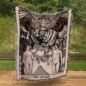 May include: A tapestry throw blanket featuring a vintage illustration of the Lovers tarot card. The card depicts a winged angel with a crown above its head, standing between a man and a woman. The text "THE LOVERS" is printed at the bottom of the card.
