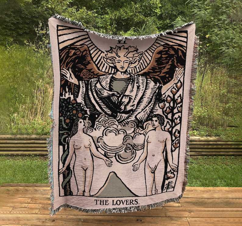 May include: A tapestry throw blanket featuring a vintage illustration of the Lovers tarot card. The card depicts a winged angel with a crown above its head, standing between a man and a woman. The text "THE LOVERS" is printed at the bottom of the card.