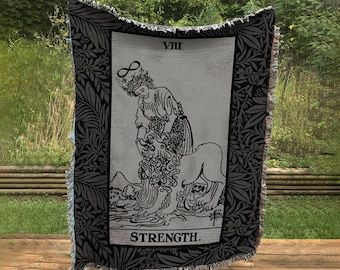 May include: Black and white woven tapestry featuring the Strength tarot card. The card depicts a woman with a crown and a halo, gently holding the mouth of a lion. The card number, VIII, and the word STRENGTH are printed in black at the top and bottom of the card.