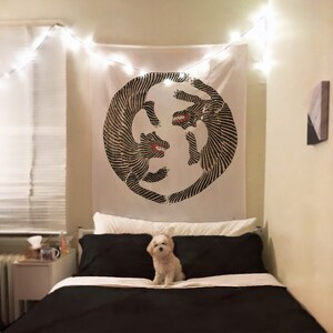 May include: A white tapestry with a black and white illustration of two stylized animals facing each other in a circle. The tapestry is hanging on a wall above a black bed with white bedding. A small white dog is sitting on the bed.