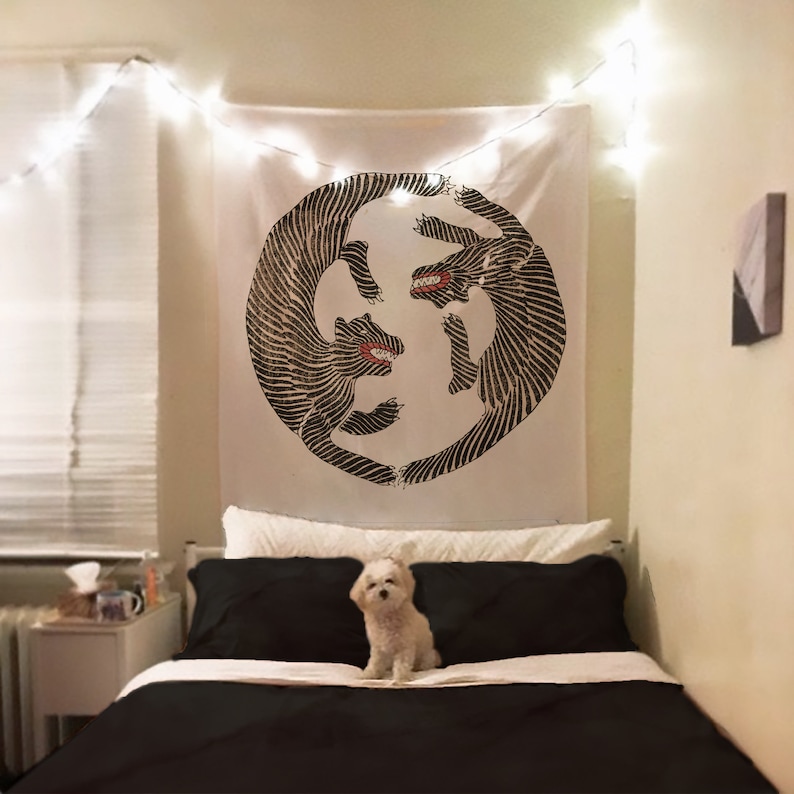 May include: A white tapestry with a black and white illustration of two stylized animals facing each other in a circle. The tapestry is hanging on a wall above a black bed with white bedding. A small white dog is sitting on the bed.