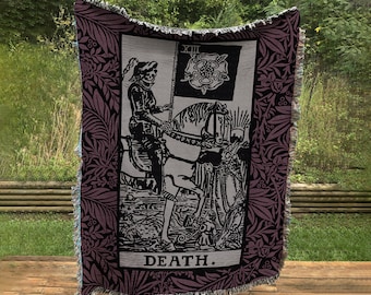 May include: A woven tapestry throw blanket with a black and white image of the Death tarot card. The card features a skeletal figure riding a white horse with a black banner that reads "XIII". The background is a dark purple with a floral pattern.