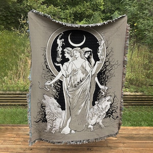 May include: A woven tapestry featuring three women standing in front of a crescent moon. The women are wearing flowing robes and have long hair. Two wolves are flanking the women, and a snake is curled around the base of the moon. The tapestry is beige with black and white details.