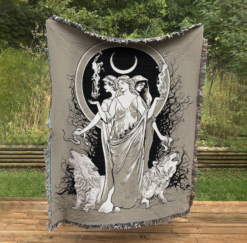 May include: A woven tapestry featuring three women standing in front of a crescent moon. The women are wearing flowing robes and have long hair. Two wolves are flanking the women, and a snake is curled around the base of the moon. The tapestry is beige with black and white details.