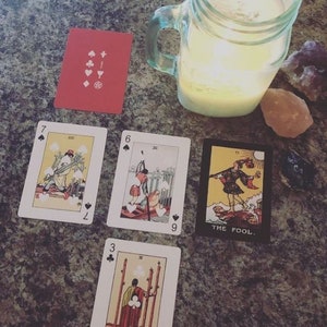 May include: Tarot card reading setup featuring playing cards with various illustrations, including "The Fool" card. A lit candle in a mason jar and crystals are also present, creating a mystical atmosphere. The cards are arranged on a textured surface.