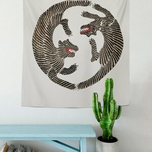 May include: A tapestry featuring a black and white illustration of two stylized animals with red teeth, facing each other in a circular design. The tapestry is hanging on a white wall above a light blue wooden bench.