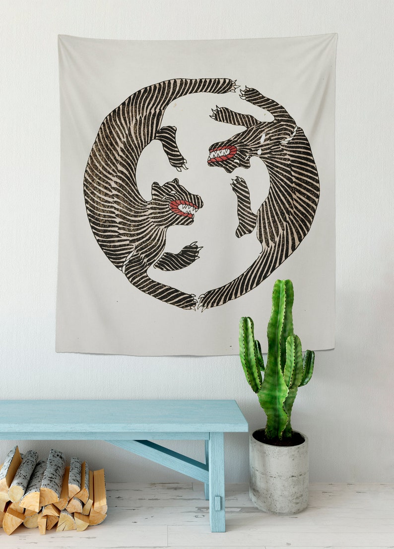 May include: A tapestry featuring a black and white illustration of two stylized animals with red teeth, facing each other in a circular design. The tapestry is hanging on a white wall above a light blue wooden bench.
