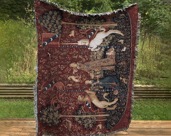 May include: A tapestry throw blanket with a red background and intricate woven designs. The tapestry features a unicorn, a knight, and a forest scene. The tapestry is fringed on all sides.