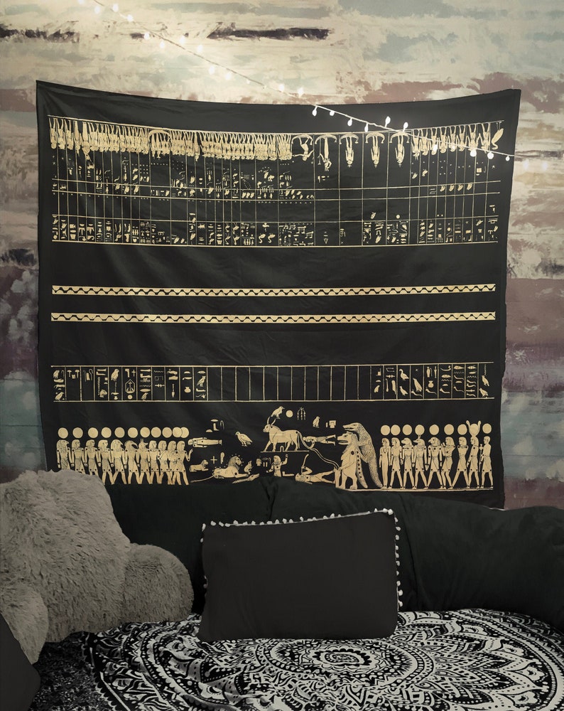 May include: Black tapestry with gold hieroglyphics and figures depicting a procession of people and animals. The tapestry is hanging on a wall with string lights.