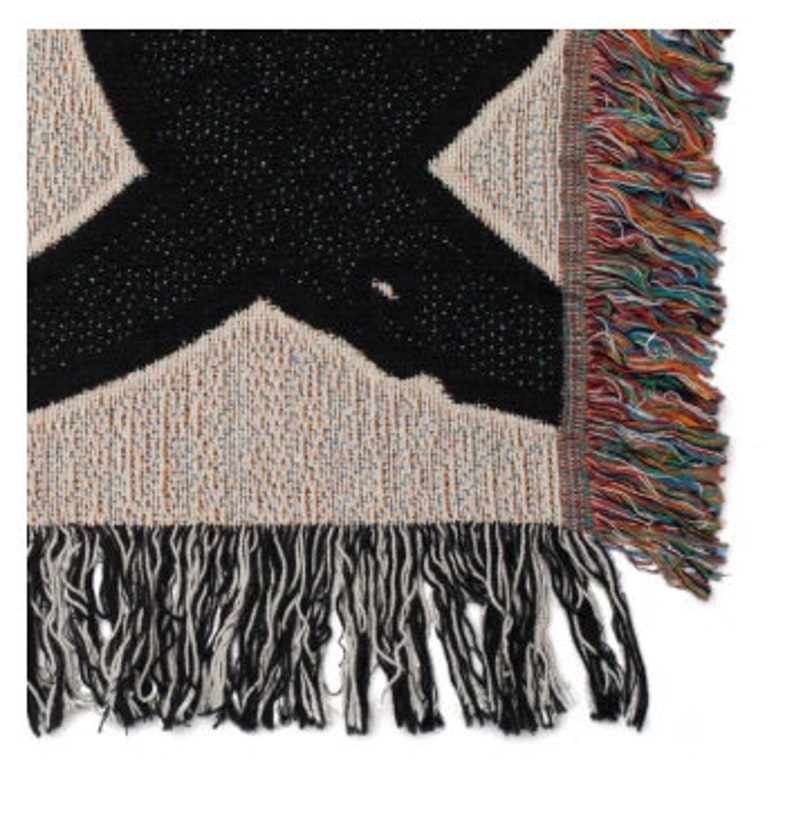May include: A woven throw blanket with a large black X design on a beige background. The blanket has a fringed edge with a mix of colorful threads. The texture of the blanket is visible, showing the weave of the fabric.