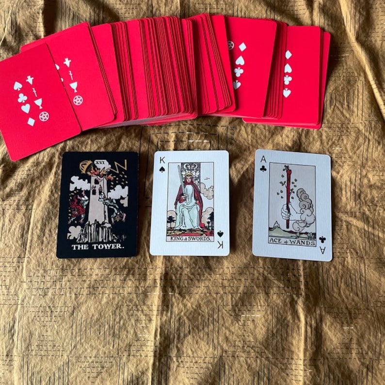 May include: A deck of tarot cards with red backs and white symbols. Three face-up cards show "The Tower," "King of Swords," and "Ace of Wands." The cards are arranged on a textured, golden-brown surface.