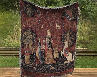 May include: A tapestry throw blanket featuring a medieval scene with a unicorn, a lion, and three women. The tapestry is woven with intricate details and vibrant colors, including red, blue, green, and gold.