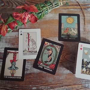May include: A collection of tarot cards featuring illustrations and text, including "The Moon," "The World," and "The Hanged Man." A bundle of green leaves and red flowers is tied with twine. The cards are arranged on a wooden surface.