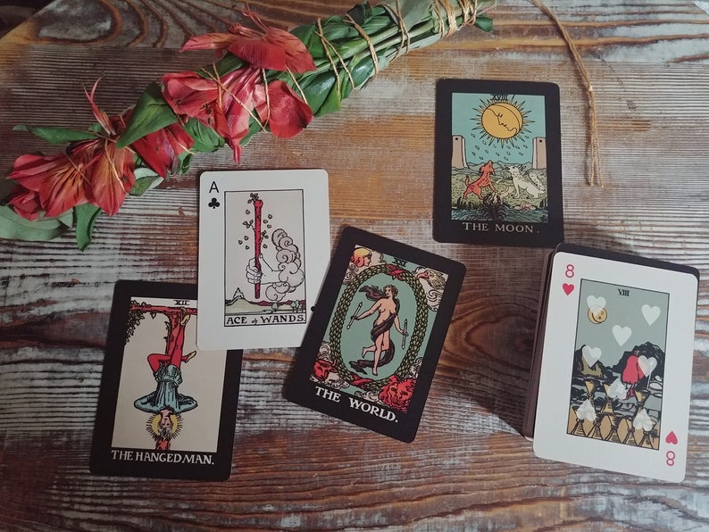 May include: A collection of tarot cards featuring illustrations and text, including "The Moon," "The World," and "The Hanged Man." A bundle of green leaves and red flowers is tied with twine. The cards are arranged on a wooden surface.