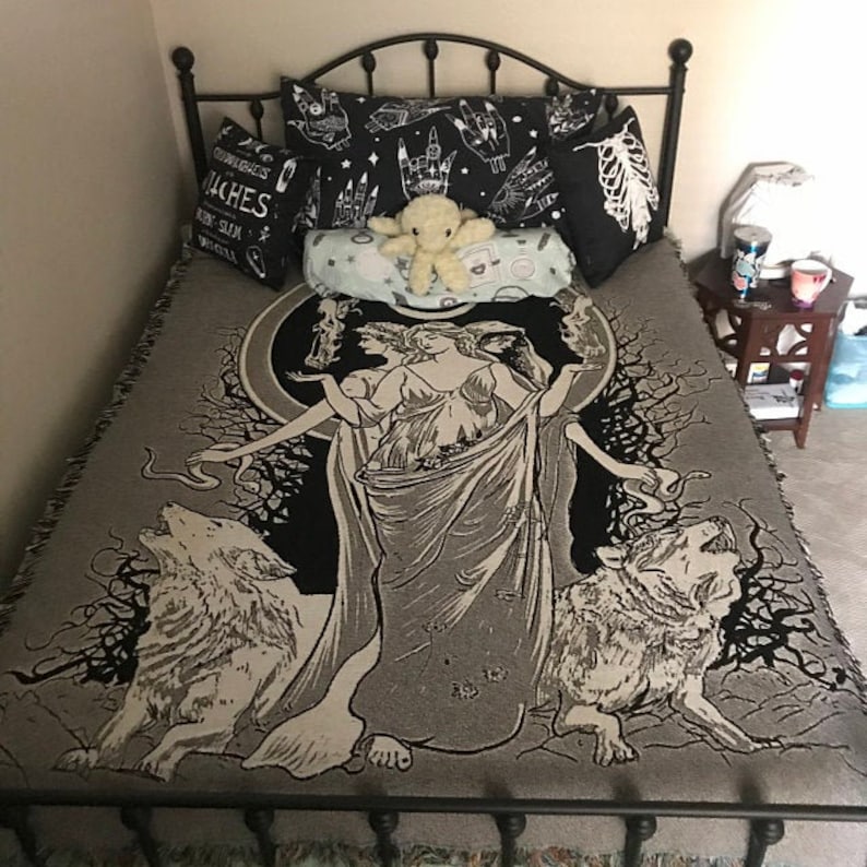 May include: A black metal bed frame with a gray and black tapestry featuring a woman with wolves and a tree. The tapestry has a black and white design with a white background. The bed has a white plush toy on it.