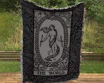 May include: Black and white woven tapestry featuring the World tarot card. The card depicts a woman standing within a wreath, holding two wands. The text "THE WORLD." is printed at the bottom.