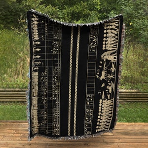 May include: Black woven throw blanket with a beige Egyptian hieroglyphic design. The blanket features vertical stripes and a fringed edge. The design includes figures, symbols, and columns of text, creating a historical theme.