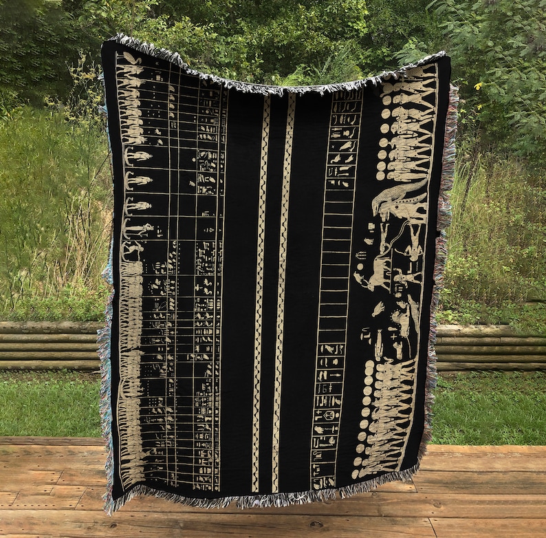 May include: Black woven throw blanket with a beige Egyptian hieroglyphic design. The blanket features vertical stripes and a fringed edge. The design includes figures, symbols, and columns of text, creating a historical theme.