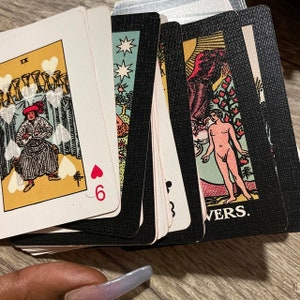 May include: A deck of tarot cards with various illustrations and symbols. The cards feature images of figures, stars, and hearts. One card shows the number 6 and a heart symbol. The cards are on a wooden surface.