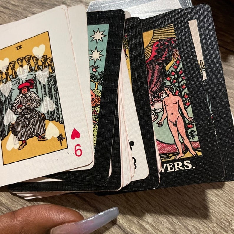 May include: A deck of tarot cards with various illustrations and symbols. The cards feature images of figures, stars, and hearts. One card shows the number 6 and a heart symbol. The cards are on a wooden surface.