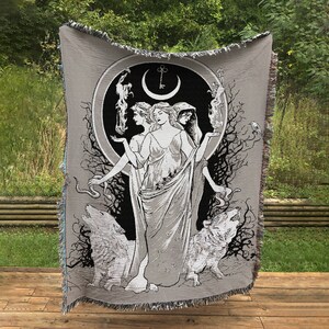 May include: A woven tapestry featuring a black and white illustration of three women standing in a circle with a crescent moon and key above them. The women are surrounded by trees and wolves. The tapestry is gray with a fringed edge.