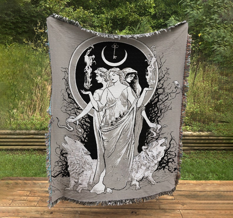 May include: A woven tapestry featuring a black and white illustration of three women standing in a circle with a crescent moon and key above them. The women are surrounded by trees and wolves. The tapestry is gray with a fringed edge.