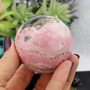 May include: A polished, spherical gemstone with a pink and white marbled pattern, accented with black and gray inclusions. The sphere is held in a hand with a dark purple nail polish. The background features green plants.