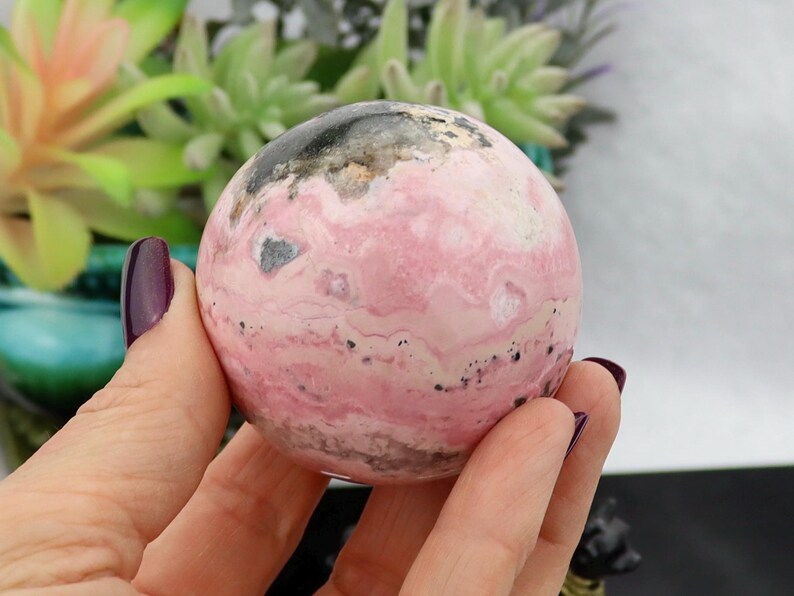 May include: A polished, spherical gemstone with a pink and white marbled pattern, accented with black and gray inclusions. The sphere is held in a hand with a dark purple nail polish. The background features green plants.