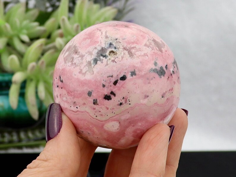 May include: A pink and white sphere with black speckles is held in a hand with a purple painted fingernail. The sphere has a marbled appearance. Green plants are visible in the background.