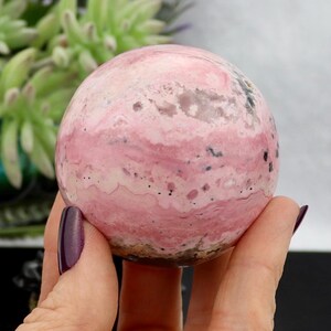 May include: A polished, spherical gemstone with a swirling pattern of pink and white hues. The stone has small black inclusions and a smooth, rounded shape. The gemstone is held in a hand with a dark purple nail polish.